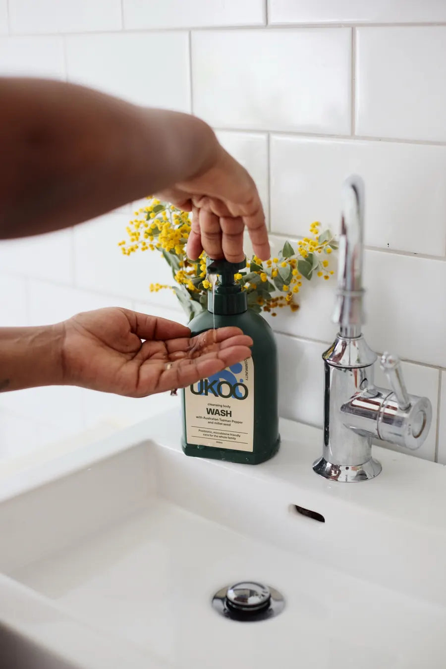 Hands under the pump of UKOO body wash beside a sprig of native wattle