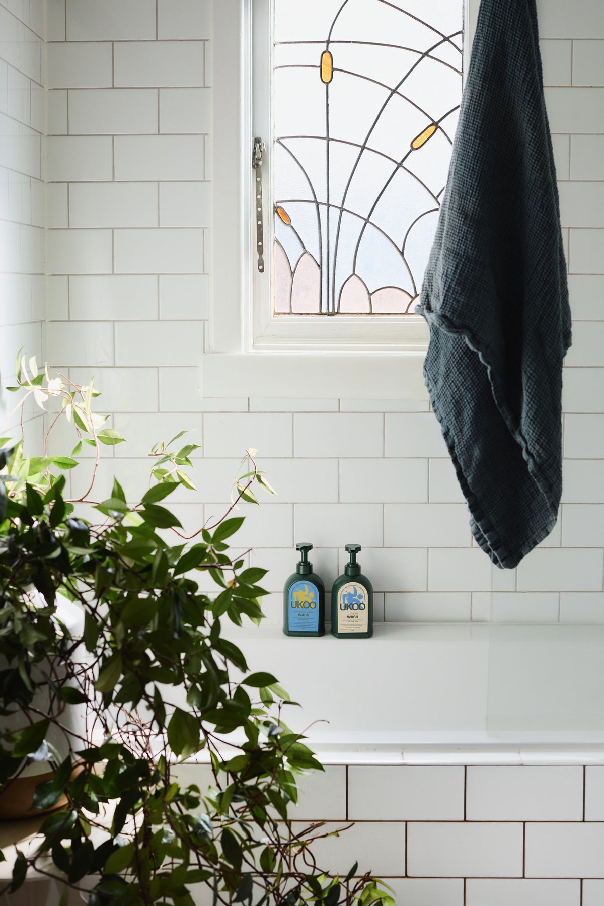 Two UKOO body washes on a white-tiled bath ledge beneath a leadlight window, a towel hanging to one side and greenery to the other