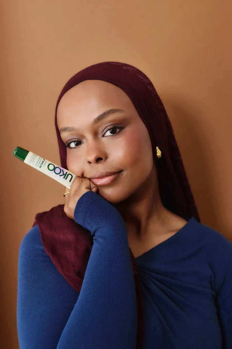 A woman in a burgundy headscarf smiles, holding a UKOO lip balm